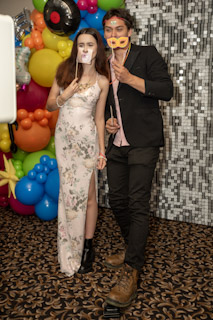 Two people pose at a photo booth with playful props, standing in front of a colorful balloon display and a silver sequin backdrop.